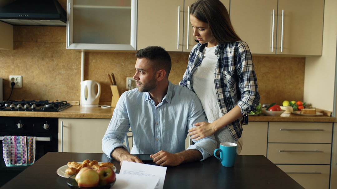 Upset young man reading unpaid bills and hugged by his wife supporting him early morning in the kitchen at home